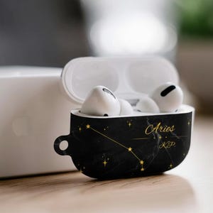 Puede incluir: Funda para AirPod negra con diseño de mármol y constelación dorada, con la palabra "Aries" en letras doradas. La funda está abierta, revelando AirPods blancos. La funda tiene un pequeño lazo para sujetar.