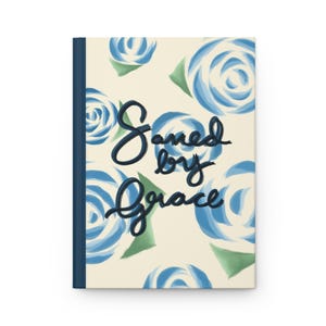 Pode incluir: Um diário de capa dura com fundo creme apresentando um padrão de rosas pintadas em azul e folhas verdes. O texto "Saved by Grace" está escrito em uma caligrafia cursiva azul-escura no centro da capa frontal.