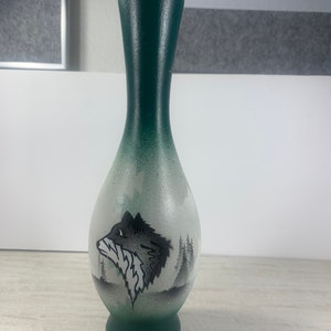 May include: A green glass vase with a white and black wolf design. The wolf is facing left and has a forest scene behind it.