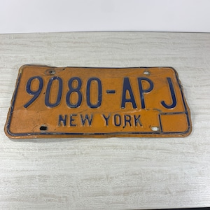 May include: Vintage New York license plate with the number 9080-AP J in dark blue against an orange background. The words "NEW YORK" are printed at the bottom. The plate shows signs of wear.