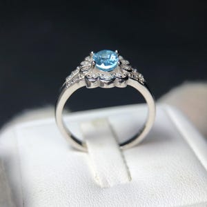 Puede incluir: Un anillo plateado con una piedra preciosa azul claro. La piedra central est&aacute; rodeada de piedras claras m&aacute;s peque&ntilde;as y un dise&ntilde;o floral. El anillo se muestra sobre una superficie blanca, sobre un fondo oscuro.
