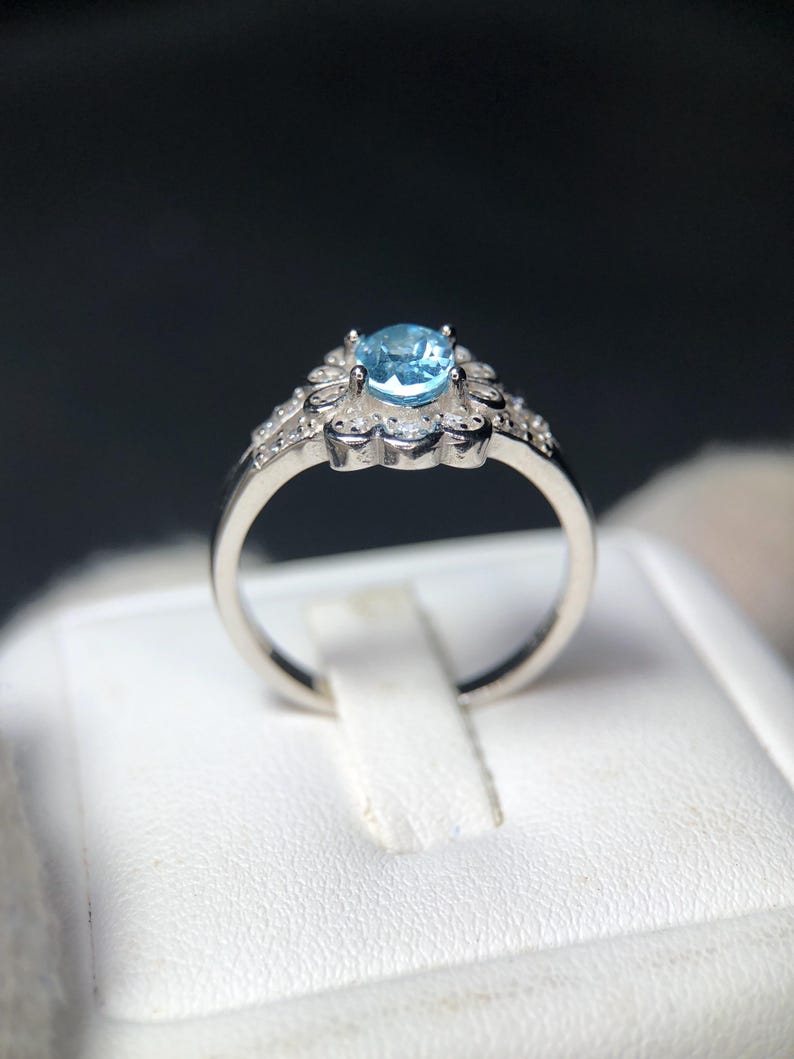 Puede incluir: Un anillo plateado con una piedra preciosa azul claro. La piedra central est&aacute; rodeada de piedras claras m&aacute;s peque&ntilde;as y un dise&ntilde;o floral. El anillo se muestra sobre una superficie blanca, sobre un fondo oscuro.