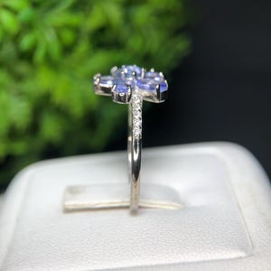 Peut inclure: Une bague en argent orn&eacute;e d'un motif floral compos&eacute; de pierres pr&eacute;cieuses ovales bleu clair. De petits diamants transparents ornent la bande. La bague est pr&eacute;sent&eacute;e sur une surface blanche devant un fond vert flou.