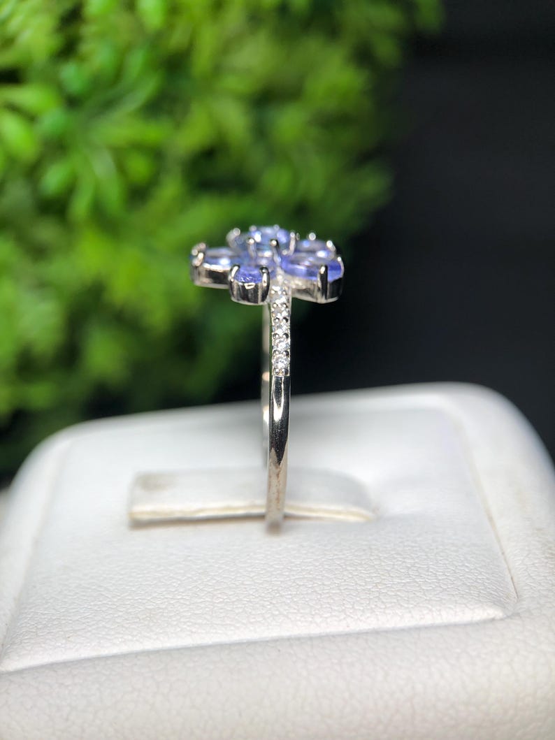 Peut inclure: Une bague en argent orn&eacute;e d'un motif floral compos&eacute; de pierres pr&eacute;cieuses ovales bleu clair. De petits diamants transparents ornent la bande. La bague est pr&eacute;sent&eacute;e sur une surface blanche devant un fond vert flou.