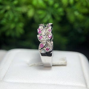 May include: A silver ring with a floral design, featuring small, round, pink gemstones. The ring is set against a blurred green background, with a white surface in the foreground.