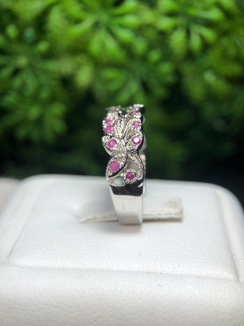 May include: A silver ring with a floral design, featuring small, round, pink gemstones. The ring is set against a blurred green background, with a white surface in the foreground.