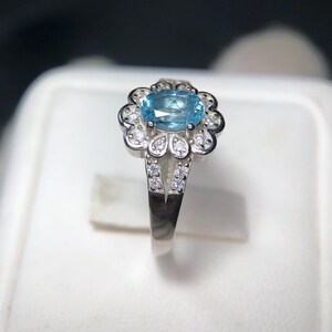 Puede incluir: Anillo de plata con una piedra preciosa ovalada azul claro, rodeada por un engaste en forma de flor. Peque&ntilde;as piedras transparentes acent&uacute;an los p&eacute;talos y la banda dividida, creando un efecto brillante. El anillo se muestra sobre un fondo blanco.