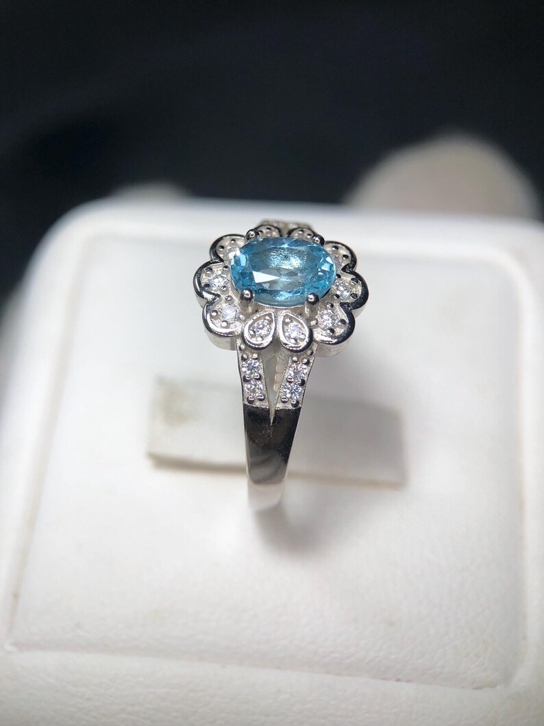 Puede incluir: Anillo de plata con una piedra preciosa ovalada azul claro, rodeada por un engaste en forma de flor. Peque&ntilde;as piedras transparentes acent&uacute;an los p&eacute;talos y la banda dividida, creando un efecto brillante. El anillo se muestra sobre un fondo blanco.