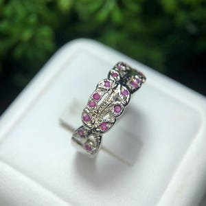 May include: A silver ring with a repeating pattern of small, round, pink gemstones. The ring has an open, interwoven design, with the gemstones set within the pattern. The ring is displayed on a white surface, with a blurred green background.