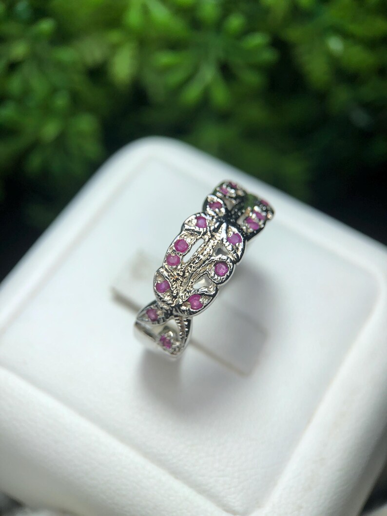 May include: A silver ring with a repeating pattern of small, round, pink gemstones. The ring has an open, interwoven design, with the gemstones set within the pattern. The ring is displayed on a white surface, with a blurred green background.