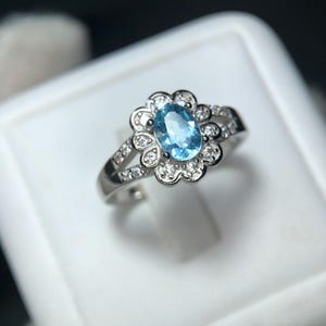 Puede incluir: Anillo de plata con una piedra preciosa ovalada azul claro en el centro, rodeada por un halo en forma de flor de piedras preciosas claras m&aacute;s peque&ntilde;as. La banda tambi&eacute;n est&aacute; adornada con peque&ntilde;as piedras preciosas claras. Sobre fondo blanco.