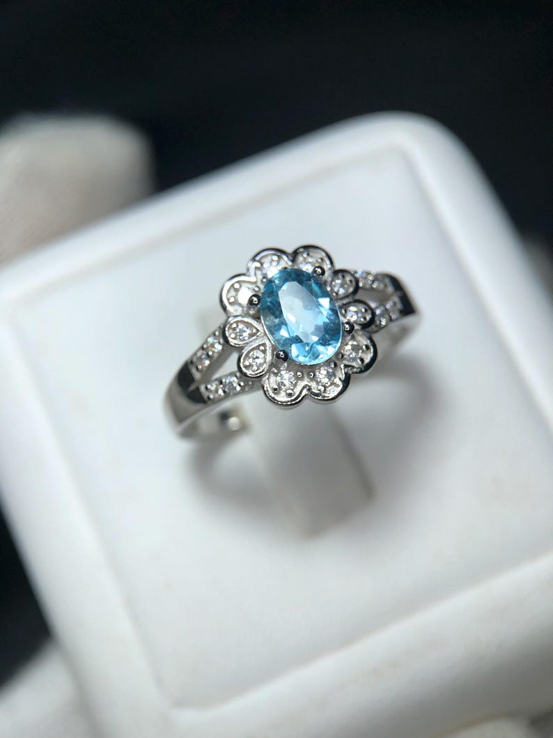 Puede incluir: Anillo de plata con una piedra preciosa ovalada azul claro en el centro, rodeada por un halo en forma de flor de piedras preciosas claras m&aacute;s peque&ntilde;as. La banda tambi&eacute;n est&aacute; adornada con peque&ntilde;as piedras preciosas claras. Sobre fondo blanco.
