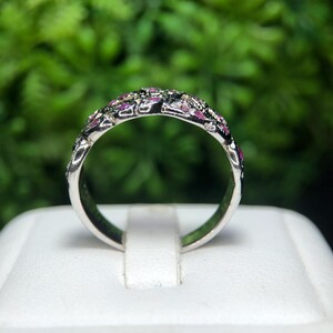 May include: A silver ring with a band adorned with small, pink gemstones and darker accents. The ring is set against a blurred green background, highlighting the intricate details of the jewelry.
