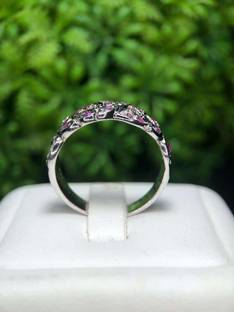 May include: A silver ring with a band adorned with small, pink gemstones and darker accents. The ring is set against a blurred green background, highlighting the intricate details of the jewelry.