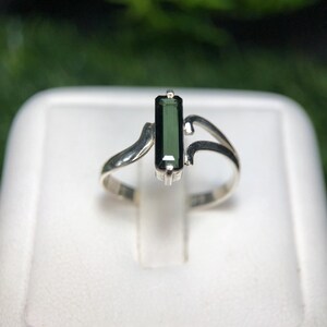 Könnte beinhalten: Ein Silberring mit einem rechteckigen, dunkelgrünen Edelstein. Der Ring hat ein einzigartiges, fließendes Design mit geschwungenen Akzenten auf beiden Seiten des Edelsteins. Das Band ist schlichtes, poliertes Silber.