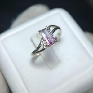 Könnte beinhalten: Ein silberner Ring mit einem rechteckigen, zweifarbigen Edelstein in Lila- und Rosatönen. Der Edelstein ist in einem silbernen Band mit einem filigranen, offenen Design an den Seiten gefasst. Der Ring wird auf einer weißen Oberfläche präsentiert.