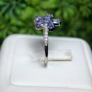 Peut inclure: Une bague en argent orn&eacute;e d'un motif floral avec des pierres pr&eacute;cieuses ovales bleu clair. La bande de la bague est orn&eacute;e de petites pierres pr&eacute;cieuses transparentes. L'arri&egrave;re-plan est vert flou.