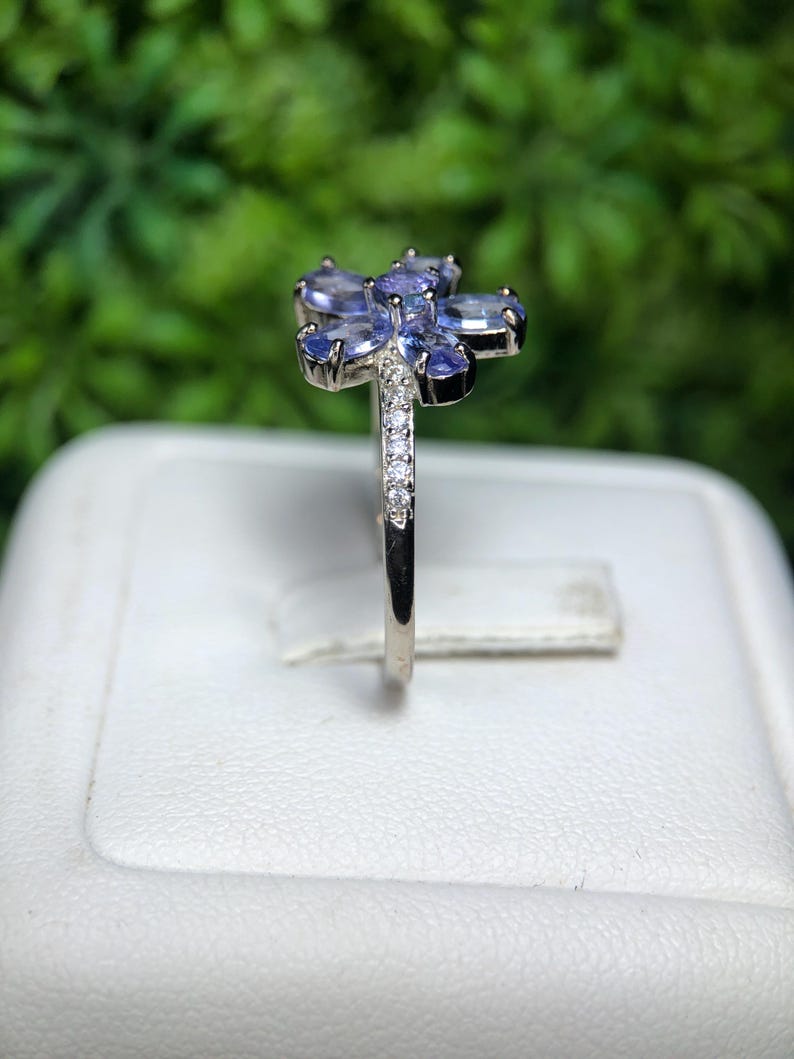 Peut inclure: Une bague en argent orn&eacute;e d'un motif floral avec des pierres pr&eacute;cieuses ovales bleu clair. La bande de la bague est orn&eacute;e de petites pierres pr&eacute;cieuses transparentes. L'arri&egrave;re-plan est vert flou.