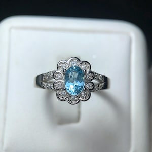 Puede incluir: Un anillo de plata con una piedra preciosa ovalada azul claro, rodeada de peque&ntilde;as piedras claras. El dise&ntilde;o del anillo incluye un engaste en forma de flor alrededor de la piedra central y piedras adicionales a lo largo de la banda.
