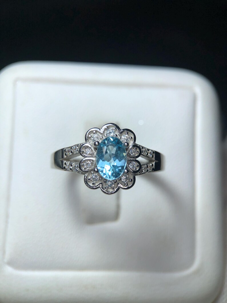 Puede incluir: Un anillo de plata con una piedra preciosa ovalada azul claro, rodeada de peque&ntilde;as piedras claras. El dise&ntilde;o del anillo incluye un engaste en forma de flor alrededor de la piedra central y piedras adicionales a lo largo de la banda.