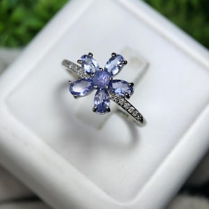 Peut inclure: Bague en argent orn&eacute;e d'un motif floral. Elle pr&eacute;sente six pierres pr&eacute;cieuses ovales bleu clair autour d'une pierre centrale bleu plus fonc&eacute;. Le corps de la bague est orn&eacute; de petits gemmes clairs.