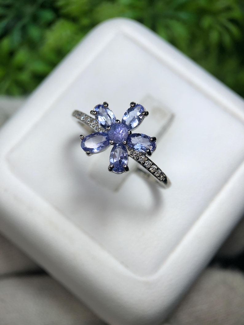 Peut inclure: Bague en argent orn&eacute;e d'un motif floral. Elle pr&eacute;sente six pierres pr&eacute;cieuses ovales bleu clair autour d'une pierre centrale bleu plus fonc&eacute;. Le corps de la bague est orn&eacute; de petits gemmes clairs.