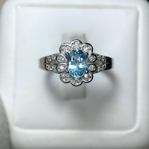 Puede incluir: Un anillo de plata con una piedra preciosa ovalada azul claro en el centro, rodeada por un halo en forma de flor de piedras preciosas claras m&aacute;s peque&ntilde;as. La banda est&aacute; detallada con piedras preciosas peque&ntilde;as adicionales.