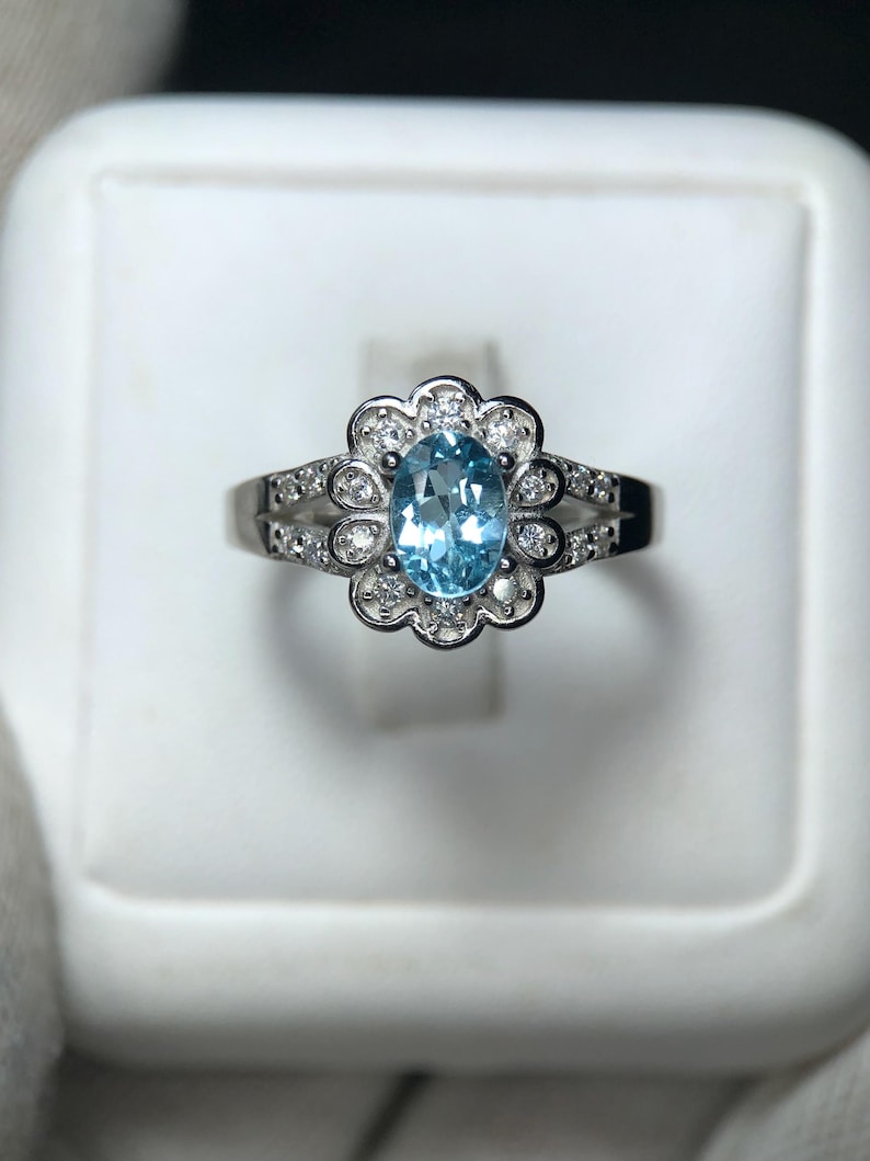 Puede incluir: Un anillo de plata con una piedra preciosa ovalada azul claro en el centro, rodeada por un halo en forma de flor de piedras preciosas claras m&aacute;s peque&ntilde;as. La banda est&aacute; detallada con piedras preciosas peque&ntilde;as adicionales.