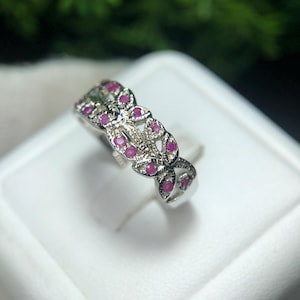 May include: A silver ring with a floral design, set with small, round, bright pink gemstones. The ring is displayed on a white surface, with a blurred green background.