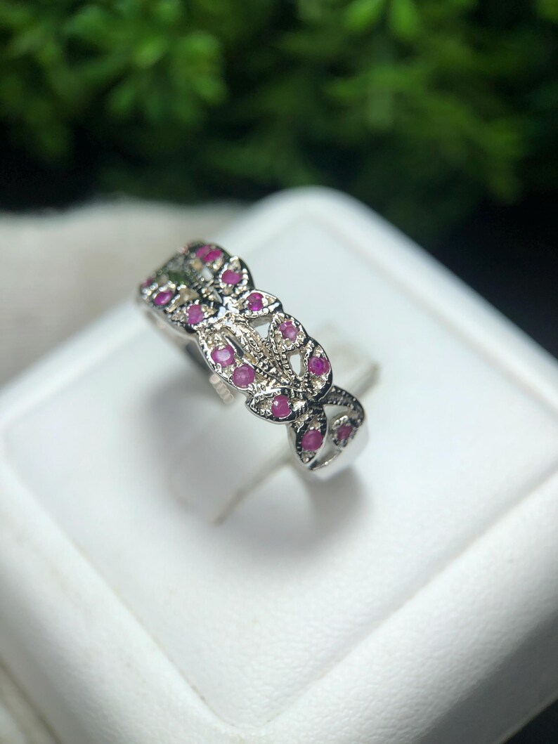 May include: A silver ring with a floral design, set with small, round, bright pink gemstones. The ring is displayed on a white surface, with a blurred green background.