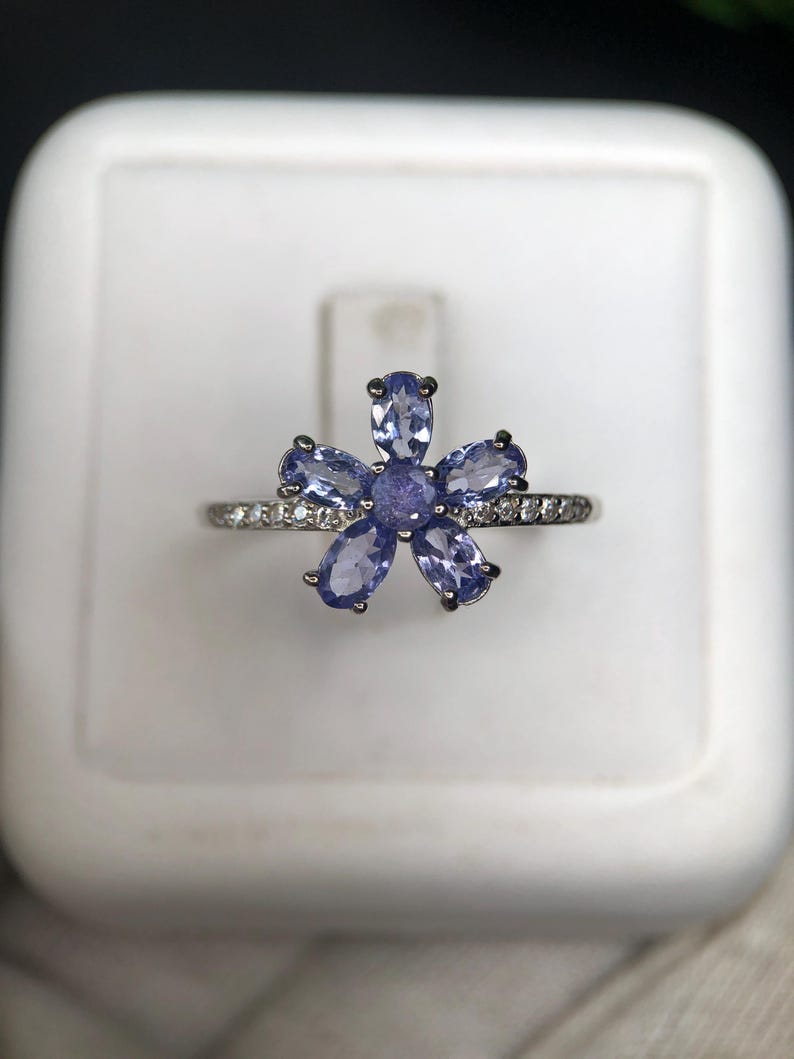 Peut inclure: Bague en argent orn&eacute;e d'une fleur compos&eacute;e de six pierres bleues claires. La monture de la bague est orn&eacute;e de petits brillants transparents. Les p&eacute;tales de la fleur sont bleu clair, sur fond blanc.