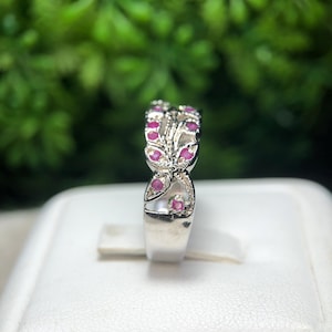 May include: A silver ring with a floral design, featuring small, round, red gemstones. The ring is set against a blurred green background, showcasing the intricate details of the jewelry.
