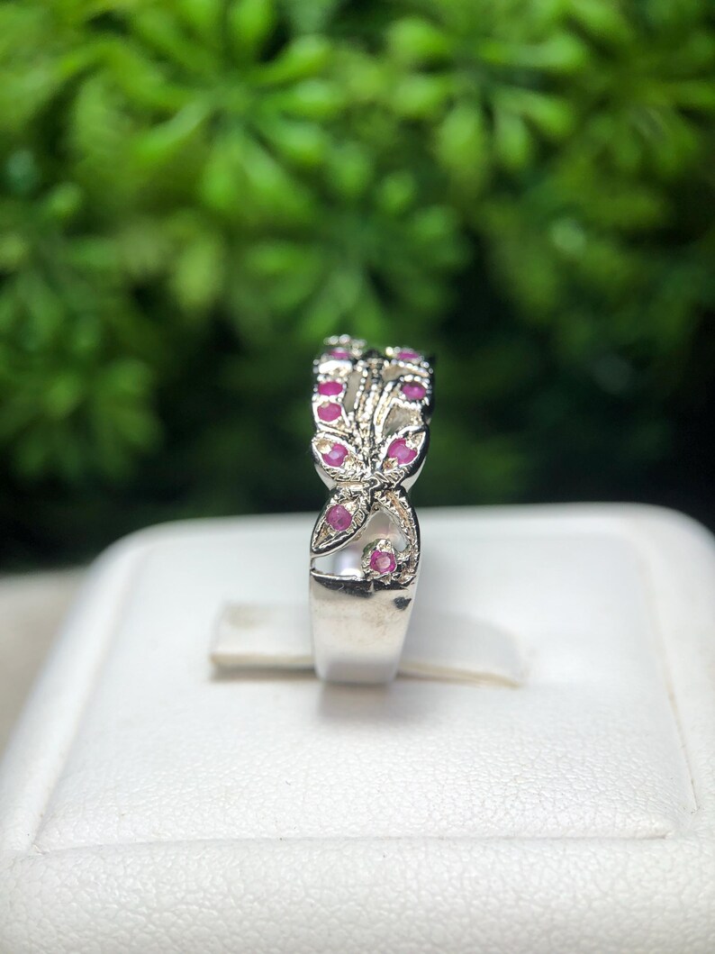 May include: A silver ring with a floral design, featuring small, round, red gemstones. The ring is set against a blurred green background, showcasing the intricate details of the jewelry.