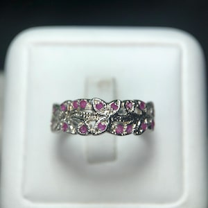 May include: A silver ring with a floral design, featuring small, round, red gemstones. The ring has an openwork pattern with a delicate, detailed appearance. The gemstones are evenly spaced throughout the design.