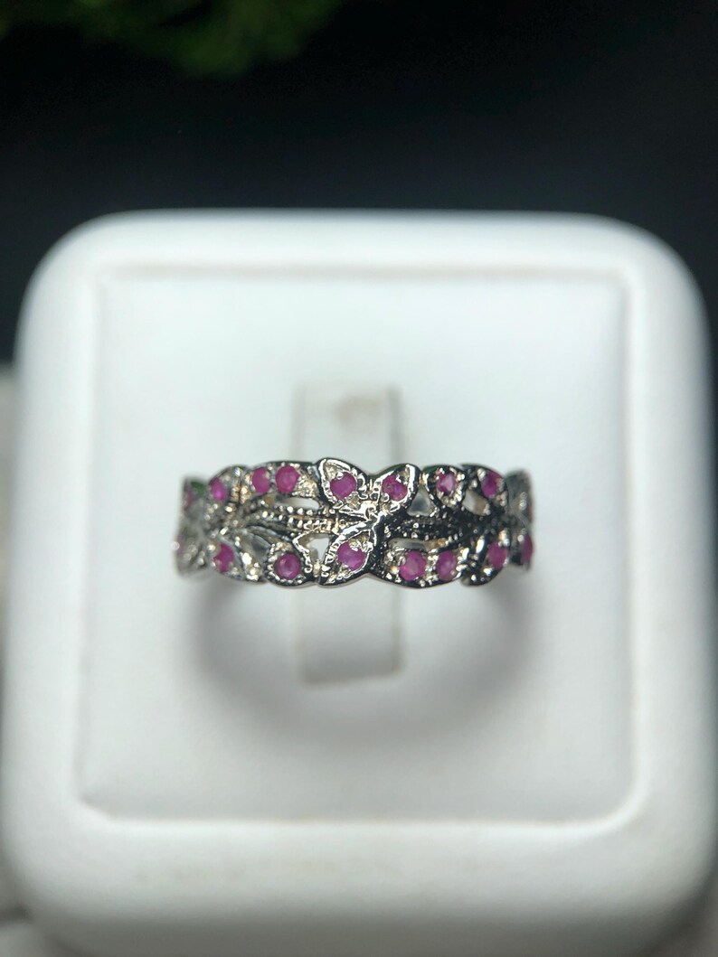 May include: A silver ring with a floral design, featuring small, round, red gemstones. The ring has an openwork pattern with a delicate, detailed appearance. The gemstones are evenly spaced throughout the design.