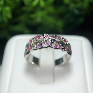 May include: A silver ring with a floral design, set with small, vibrant red gemstones. The ring is displayed on a white surface, with a blurred green background. The gemstones are evenly spaced, creating a continuous pattern around the band.