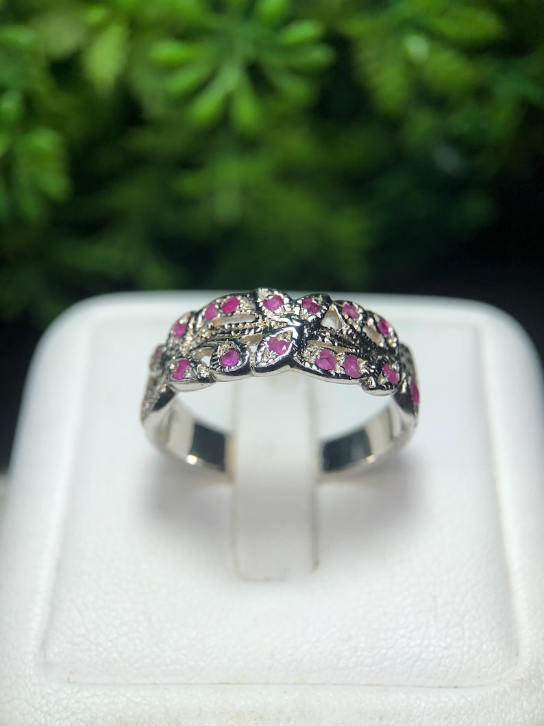 May include: A silver ring with a floral design, set with small, vibrant red gemstones. The ring is displayed on a white surface, with a blurred green background. The gemstones are evenly spaced, creating a continuous pattern around the band.