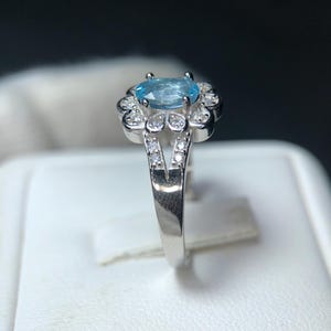 Puede incluir: Un anillo de plata con una piedra preciosa ovalada de color azul claro, rodeada de peque&ntilde;as piedras preciosas transparentes. La banda del anillo se divide en forma de V, con piedras preciosas transparentes adicionales en los lados. El anillo se muestra sobre una superficie blanca.