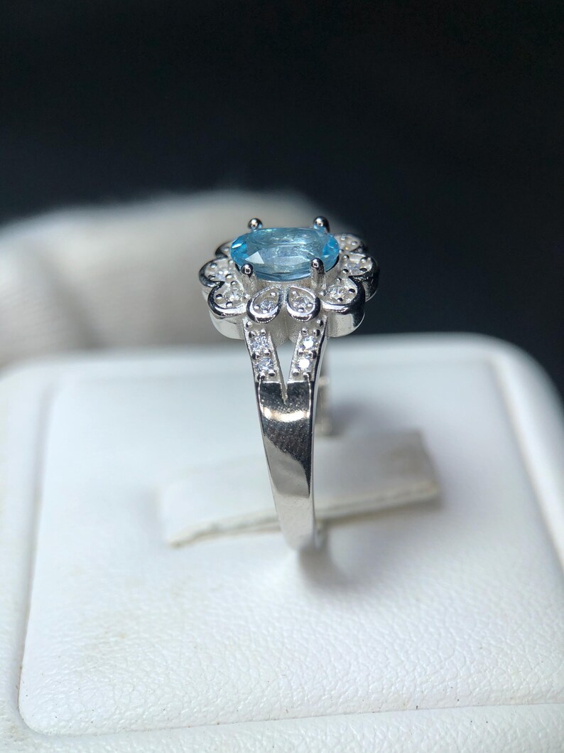 Puede incluir: Un anillo de plata con una piedra preciosa ovalada de color azul claro, rodeada de peque&ntilde;as piedras preciosas transparentes. La banda del anillo se divide en forma de V, con piedras preciosas transparentes adicionales en los lados. El anillo se muestra sobre una superficie blanca.