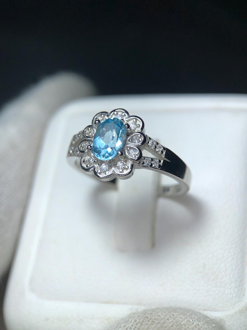 Puede incluir: Un anillo de plata con una piedra preciosa ovalada azul claro, rodeada por un dise&ntilde;o floral con peque&ntilde;as piedras de acento claras. La banda del anillo tambi&eacute;n presenta peque&ntilde;as piedras.