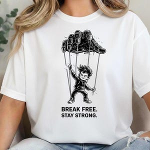 May include: White t-shirt featuring a black and white graphic of a hand controlling a puppet. The puppet is a cartoon boy. The text "BREAK FREE. STAY STRONG." is below the graphic. The shirt is made of a soft material.