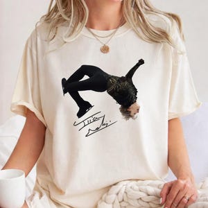 May include: Cream-colored t-shirt featuring a graphic of a figure skater in a black costume mid-performance. The skater is upside down with arms extended. Below the image is a signature in black ink. The shirt is a casual style.