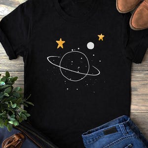 May include: Black t-shirt featuring a white planet design with a ring, white dots, two yellow stars, and a white moon. The shirt is paired with blue jeans and brown shoes on a wooden surface.
