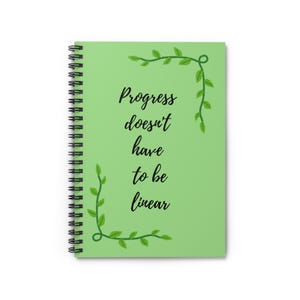 May include: A spiral-bound notebook with a light green cover. The cover features the text "Progress doesn't have to be linear" in black script, with green leaf accents. The notebook is designed for writing and note-taking.
