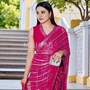 May include: A woman in a fuchsia saree with silver detailing and a matching sleeveless top. The saree is draped over the shoulder and has a geometric design. She carries a small, silver clutch. The text "MISHAB" is visible.