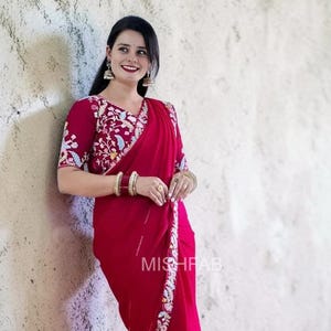 May include: A vibrant red saree with intricate floral embroidery on the sleeves and border. The saree features a matching blouse with detailed embroidery. The image shows the text "MISHFAB".