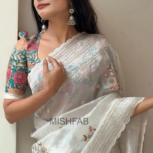 May include: A white saree with floral embroidery and a scalloped edge. The blouse has colourful floral sleeves. The text "MISHFAB" is visible on the saree. The saree is a traditional garment.