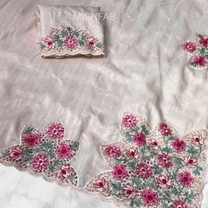 Pode incluir: Tecido branco sujo com bordados florais intrincados em rosa e verde. O design apresenta uma borda ondulada e detalhes delicados. Um quadrado de tecido dobrado correspondente também é visível.