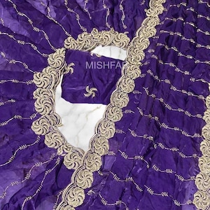 Könnte beinhalten: Ein lebendiger violetter Stoff mit aufwendiger goldener Stickerei. Das Design zeigt ein sich wiederholendes Muster aus wirbelnden floralen Motiven und zarten linearen Akzenten. Der Text "MISHFA" ist sichtbar. Dieser Stoff wird wahrscheinlich für Kleidung oder dekorative Zwecke verwendet.