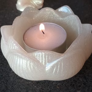 以下が含まれることがあります： 白くキラキラ光る蓮の花の形をしたキャンドルホルダーで、中に火の灯ったティーライトキャンドルが入っています。キャンドルホルダーは質感のある表面で、半透明の素材で作られています。炎は明るく輝いています。
